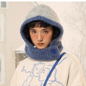 Könnte beinhalten: Eine Kapuzenschal-Kombination aus Strick in Blau und Grau. Die Kapuze ist grau mit blauem Besatz und einem blauen Knopfverschluss. Der Schal wird über einem weißen Pullover mit blauer Illustration getragen.