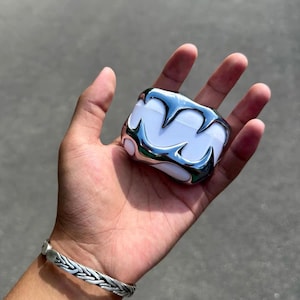 May include: A white rectangular wireless earbud charging case featuring a metallic silver overlay with sharp, pointed, claw-like protrusions extending across the front surface, creating a distinct, high-contrast, reflective, and textured appearance.