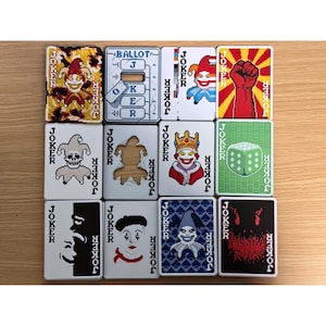 Puede incluir: Una colección de doce cartas de juego cuadradas con varios diseños de comodín. Cada carta tiene la palabra "JOKER" impresa en los lados. Los diseños incluyen un bufón, una calavera, una urna y un puño.