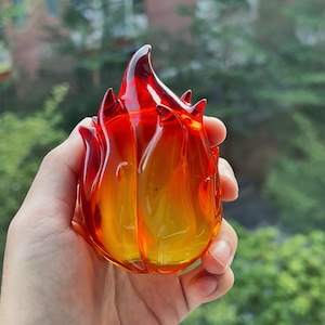 May include: A translucent glass sculpture shaped like a stylized flame, featuring a vibrant color gradient that transitions from a deep, saturated red at the pointed top down to a bright, warm yellow at the rounded base, with smooth, curved edges.