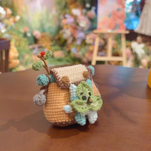 May include: A small, crocheted backpack in tan with a cream-colored flap, featuring two brown circular buttons. A green, leaf-shaped creature with a small face is attached to the front. A brown stem with a green leaf and red berry sits on top.