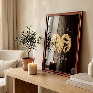 May include: A framed artwork featuring a gold ram zodiac symbol on a black background. The artwork is displayed on a light wood console table, alongside a potted plant, a lit candle, and a stack of books. The frame is a dark brown color.