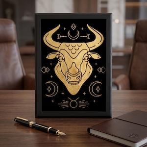 May include: Framed artwork featuring a gold Taurus zodiac symbol on a black background. The bull illustration is surrounded by celestial designs. A black pen and a brown notebook are on a wooden desk.