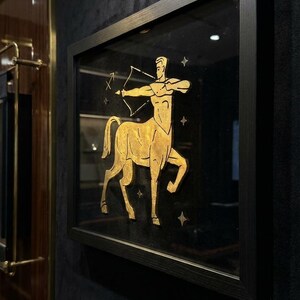 May include: Framed artwork featuring a gold-colored Sagittarius zodiac symbol on a black background. The centaur archer is depicted with a bow and arrow, surrounded by small star accents. The artwork is encased in a black frame and mounted on a dark wall.