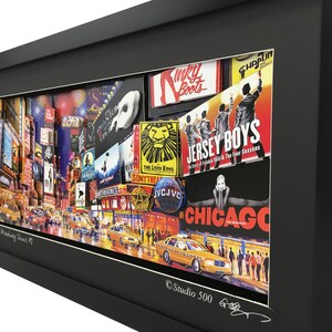 May include: A framed collage of Broadway show posters and images of Times Square, New York City. The collage features posters for shows like "The Lion King", "Chicago", "Jersey Boys", and "Kinky Boots". The collage is framed in black and has a white mat. The bottom of the frame has the text "Times Square Broadway Dreams #1" and "Studio 500".