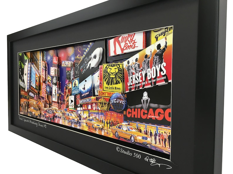 May include: A framed collage of Broadway show posters and images of Times Square, New York City. The collage features posters for shows like "The Lion King", "Chicago", "Jersey Boys", and "Kinky Boots". The collage is framed in black and has a white mat. The bottom of the frame has the text "Times Square Broadway Dreams #1" and "Studio 500".