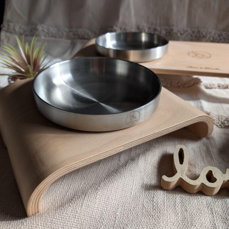May include: Two stainless steel pet bowls on a raised wooden platform. The platform is made of light wood and has a curved design. The bowls are elevated to make it easier for pets to eat and drink.