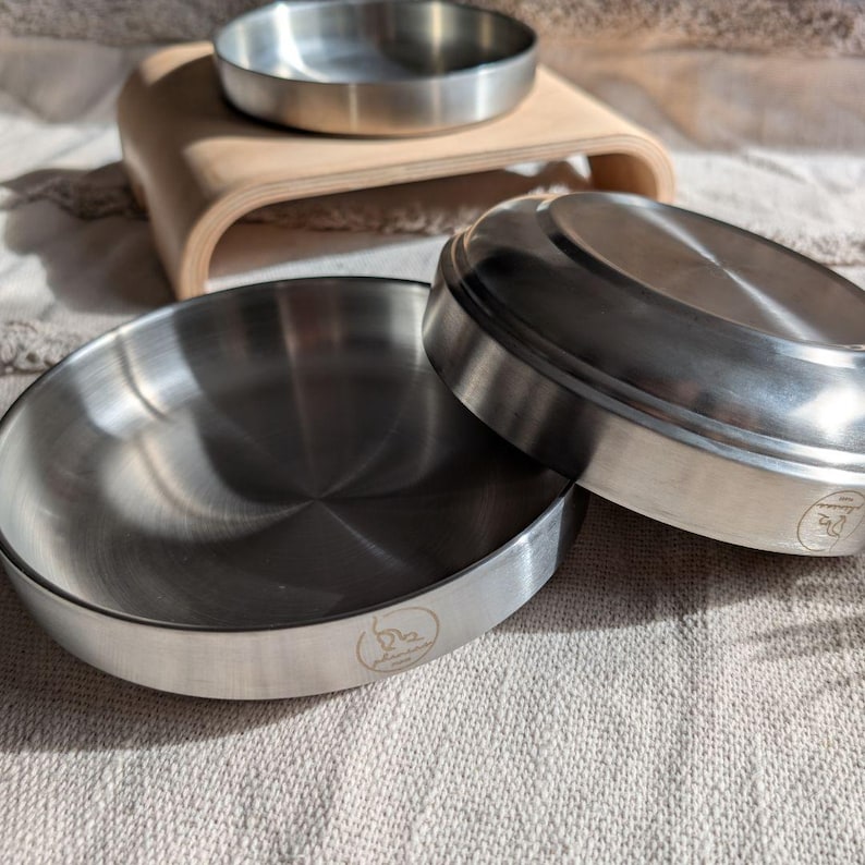 May include: Two stainless steel pet bowls, one stacked on top of the other, on a light brown wooden surface. The bowls are round and have a shiny finish.
