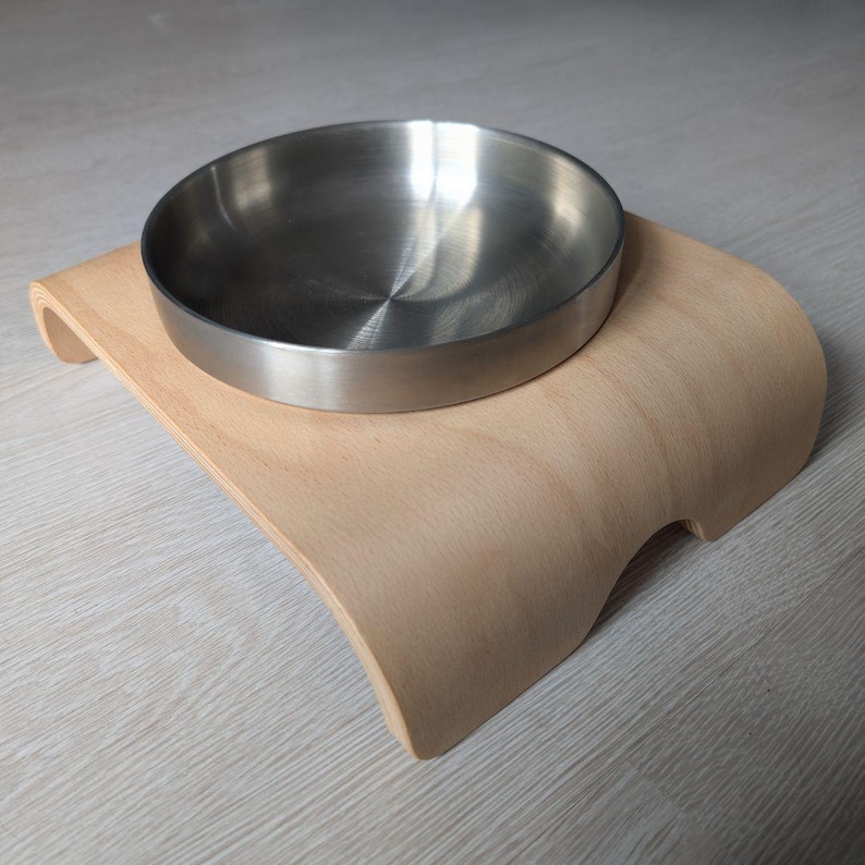 May include: A stainless steel pet bowl sits on a light brown wooden stand. The stand is curved and has a raised edge around the bowl.