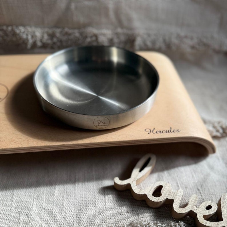 May include: A stainless steel bowl with a rounded edge sits on a wooden tray. The tray is engraved with the word "Hercules".