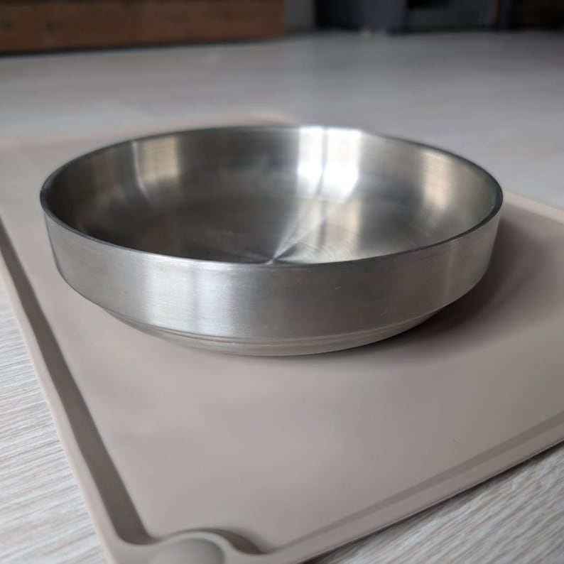 May include: A stainless steel pet bowl with a smooth, shiny finish. The bowl is sitting on a light brown silicone mat.