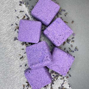 May include: Six purple bath bombs arranged in a pyramid shape on a white fabric background with dried lavender flowers scattered around them.