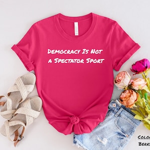 Può includere: Una maglietta rosa con il testo "Democracy is not a spectator sport" in lettere bianche. La maglietta è mostrata su uno sfondo bianco con un paio di sandali e un paio di pantaloncini di jeans.