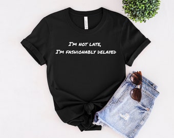 May include: Black t-shirt with white text that reads "I'm not late, I'm fashionably delayed". The shirt is folded and displayed on a white background with a pair of denim shorts and sunglasses.