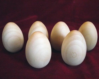 May include: Six smooth, wooden eggs in various shades of light brown, arranged on a burgundy surface. The eggs are all slightly different in size and shape, giving them a natural, handcrafted look.