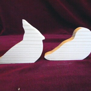 Puede incluir: Dos formas de p&aacute;jaros de madera sin terminar, un cardenal y otro p&aacute;jaro m&aacute;s peque&ntilde;o, sobre un fondo burdeos.