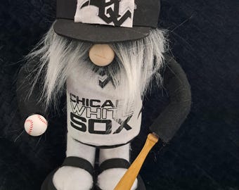 May include: A handcrafted gnome figurine wearing a black baseball cap with a white "S" logo and a white jersey with "CHICAGO WHITE SOX" printed on it. The gnome holds a baseball and a wooden bat. The base is black.