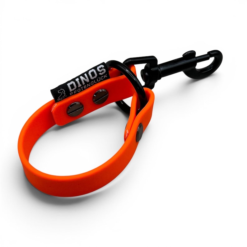 May include: A bright orange loop strap featuring a black metal swivel snap hook and a black metal ring. A black fabric tag is attached to the strap with the white text DINOS PFOTENGL&Uuml;CK and a small dinosaur icon. The strap is secured with black rivets.