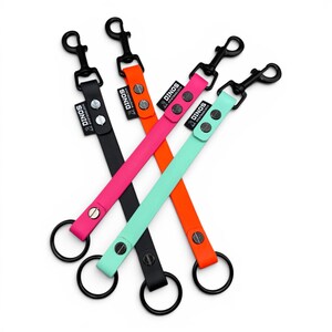 May include: Four short dog leashes in black, orange, hot pink, and mint green. Each strap features a black metal swivel snap hook at one end, a black metal O-ring at the other, and a black fabric tag labeled DINOS PFOTENGL&Uuml;CK with silver rivets.