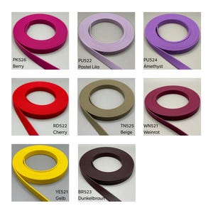 May include: A collection of eight coiled strips of flat, flexible material in various colors, each labeled with a code and color name: PK526 Berry, PU522 Pastel Lila, PU524 Amethyst, RD522 Cherry, TN525 Beige, WN521 Weinrot, YE521 Gelb, and BR523 Dunkelbraun.