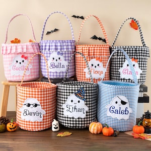 May include: A collection of seven Halloween-themed gingham fabric baskets in various colors, each with a white ghost applique and a personalized name. The baskets have handles and are decorated with bats and fall foliage.
