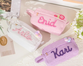 May include: Three corduroy pencil cases in white, pink, and lavender. Each case has a name embroidered on the front: Arlo, Enid, and Kari. The pink case has a lollipop applique. All have a zipper and a handle.