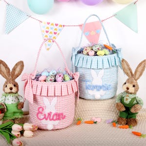 May include: Two Easter baskets, one pink and one blue, filled with decorative eggs and straw. Each basket has a gingham pattern and a bunny applique with a name. Bunny figures and carrot decorations complete the scene.