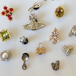 Op de afbeelding: Een verzameling decoratieve schoenbedels in verschillende ontwerpen. Bedels zijn onder meer een kruis met rode accenten, een vork met tekst, een ingelijste foto met parels, een kerstboom, een hart en een slot. De bedels zijn in goud, zilver en parelkleuren.