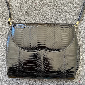 May include: A black handbag with a glossy, textured finish and a flap closure. The bag has a long, thin strap and gold-coloured hardware. The surface has a snakeskin pattern, adding a touch of luxury. The bag is rectangular in shape.