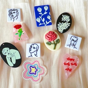 May include: A collection of ten assorted pendants featuring various shapes and designs, including line art faces on white squares, a blue rectangle with white flowers, a pink heart with red text, and oval pieces with floral and figure motifs.
