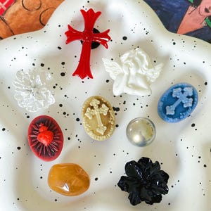 May include: A white ceramic dish with black polka dots, featuring a collection of vintage buttons. The buttons are in various shapes and colours, including a red cross, a white angel, a black flower, a yellow stone, a red heart, a blue cross, and a white cross.
