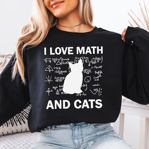 May include: Black sweatshirt with white text that reads "I LOVE MATH" and a white silhouette of a cat sitting on a background of mathematical equations. The text "AND CATS" is below the cat.