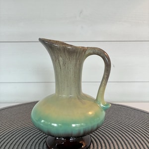 Może przedstawiać: Ceramiczny dzbanek o pękatym korpusie, rozszerzanym wylewie i uchwycie. Szkliwo przechodzi od ciemnobrązowej podstawy przez żywy turkusowy środek, aż po jasną zieleń i brązowe zacieki w górnej części naczynia przy otworze wylewowym.