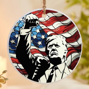 May include: A round ceramic ornament featuring a black and white illustration of a person with their fist raised in the air. The background is a red, white, and blue American flag.