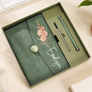 May include: A sage green notebook set in a matching gift box, featuring a floral print and the name Evelyn in white script. The notebook has a round button closure and is accompanied by a matching green pen and a silver ink refill cartridge.