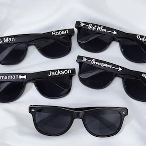 Może przedstawiać: Czarne okulary przeciwsłoneczne z białym napisem "Best Man", "Groomsman" i imionami. Okulary mają klasyczny design z ciemnymi soczewkami. Na zdjęciu kilka par, idealnych na przyjęcie weselne.
