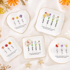 Könnte beinhalten: Eine Sammlung weißer Keramik-Schmuckschalen in verschiedenen Formen, darunter Herz, Quadrat und rund. Jede Schale zeigt ein Blumendesign mit Namen und dem Text "Sisters are different flowers from the same garden". Goldfarbene Ränder.