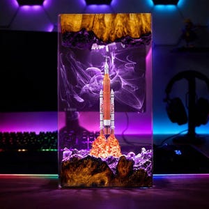 Puede incluir: Una pieza de arte de resina rectangular que representa el lanzamiento de un cohete. El cohete es naranja y blanco, con el logotipo de la NASA. La base es marrón y morada, con un resplandor naranja ardiente que emana de la base del cohete. El fondo tiene patrones morados similares al humo.