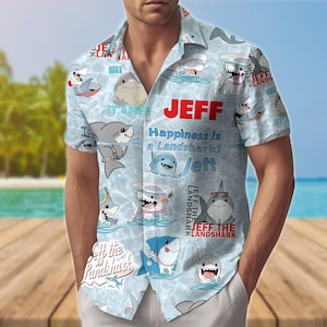 May include: Light blue button-down shirt with a shark pattern. The shirt features cartoon sharks, the name "Jeff", and the phrase "Happiness is a Landshark!" in red and blue text. The shirt has short sleeves.