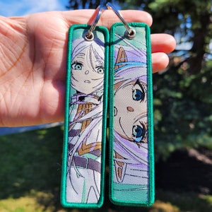 May include: Two green keychains featuring white embroidered anime characters. The characters have long white hair and are wearing white and green outfits. The keychains are approximately 7.5 cm long.