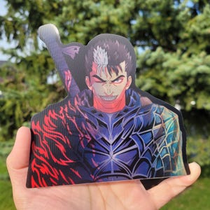 May include: A sticker featuring a character from the anime series Berserk. The character is a man with black hair and a white streak in his hair. He is wearing black armor and has a menacing expression on his face. The sticker is a holographic design with a red and blue color scheme.