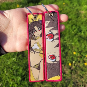 Peut inclure: Deux porte-clés avec une bordure rose. Le porte-clés de gauche représente un personnage d'anime avec de longs cheveux bruns et des yeux rouges. Le porte-clés de droite représente un gros plan d'un œil avec des détails rouges et noirs.