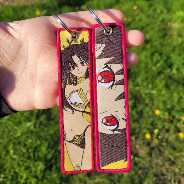 Puede incluir: Dos llaveros con un borde rosa. El llavero de la izquierda presenta un personaje de anime con cabello largo y castaño y ojos rojos. El llavero de la derecha presenta un primer plano de un ojo con detalles rojos y negros.