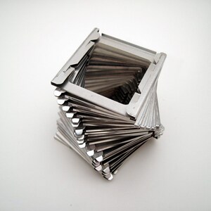 Pu&ograve; includere: Una scultura geometrica color argento con un design a spirale. La scultura &egrave; composta da molti sottili pezzi di metallo rettangolari impilati e attorcigliati per formare una forma quadrata. L'oggetto &egrave; su sfondo bianco.
