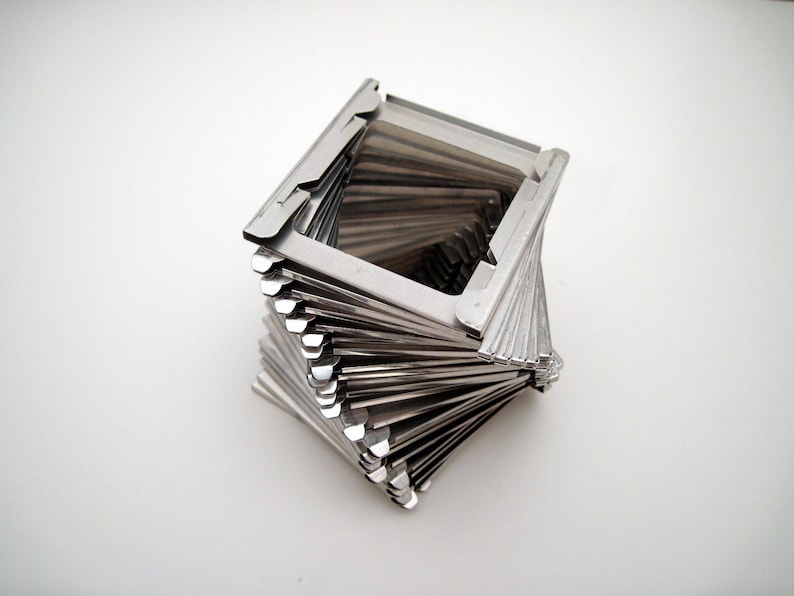 Pu&ograve; includere: Una scultura geometrica color argento con un design a spirale. La scultura &egrave; composta da molti sottili pezzi di metallo rettangolari impilati e attorcigliati per formare una forma quadrata. L'oggetto &egrave; su sfondo bianco.