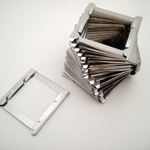 May include: A collection of silver-colored metal slide mounts, one detached and the others stacked in a spiral formation. The mounts have a rectangular shape with a clear window in the center, and are likely for holding photographic slides.