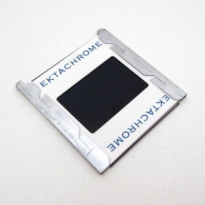 May include: A vintage Ektachrome slide mount with a black rectangular opening. The white frame has the word "Ektachrome" printed in blue. The mount is held together with silver metal clips.