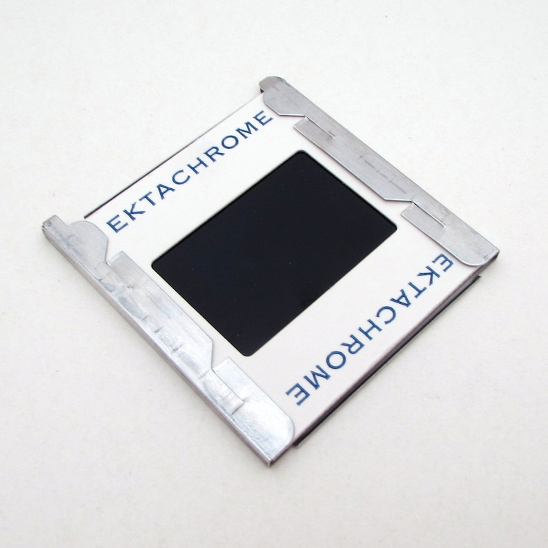 Pu&ograve; includere: Un supporto per diapositive Ektachrome vintage con un'apertura rettangolare nera. La cornice bianca ha la scritta "Ektachrome" stampata in blu. Il supporto &egrave; tenuto insieme da clip metalliche argentate.
