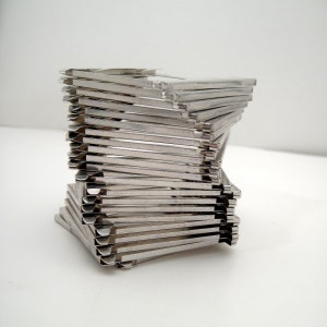 May include: A stack of silver metal clips arranged in a spiral shape. The clips are rectangular with a small folded edge on one side. The arrangement creates a three-dimensional, geometric design against a white background.