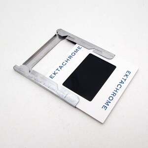 Pu&ograve; includere: Un supporto per diapositive bianco con la scritta "Ektachrome" in blu, che contiene una diapositiva nera. Il supporto &egrave; tenuto in posizione da un supporto per diapositive in metallo argentato.